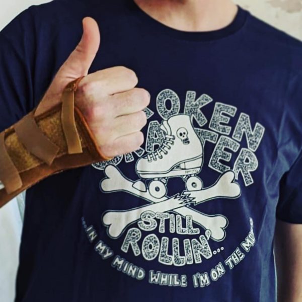 Man with Broken arm in a splint shows thumbs up wearing a navy Blue Broken skater T-shirt with roller skater and broken crossed bones.